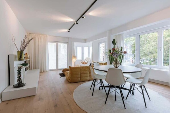 A modern living room with a dining area, large windows, and a view of greenery outside.