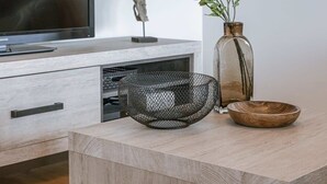 Een moderne woonkamer met een flatscreen televisie, een houten salontafel, een draadmand, een vaas met bloemen en een decoratief object op de tv-meubel.