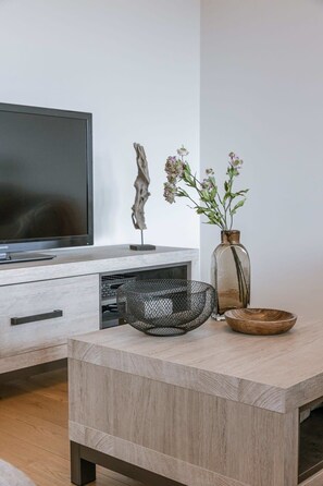 Ein modernes Wohnzimmer mit Flachbildfernseher, Couchtisch aus Holz, Drahtkorb, Blumenvase und einem dekorativen Gegenstand auf der TV-Bank.