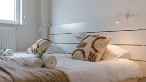 Een netjes opgemaakt bed met opgerolde handdoeken en een plaid, een bedlampje en een raam met zonwering.