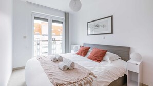Un dormitorio con una cama grande, una mesita de noche, una lámpara colgante, un cuadro enmarcado y una ventana con vistas a edificios.