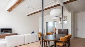 Una sala de estar moderna con sofá, mesa de comedor y sillas, suelo de madera y una lámpara colgante grande.