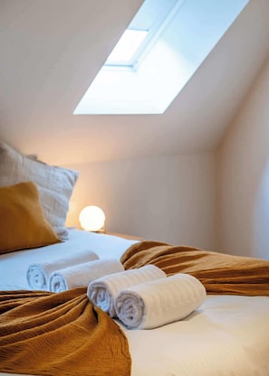 Une chambre avec un puits de lumière, un lit avec du linge de lit blanc, une couverture marron et une lampe de chevet.