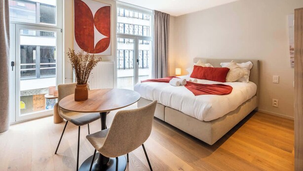 Ein modernes Hotelzimmer mit Bett, Esstisch mit Stühlen, einer Vase mit Trockenblumen und einem großen Fenster mit Vorhängen.