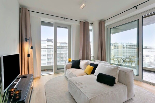 A modern living room with a sofa, a TV, and a balcony with a view of buildings. - Bright coastal retreat w/ kitchen & parking (Oostende)
