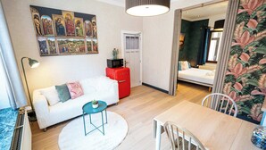 Ein modernes Hotelzimmer mit einem weißen Sofa, einem Esstisch, einem roten Koffer, einem Bild an der Wand und tropischem Wandpapier an einer Seite.