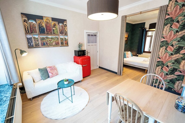 A modern hotel room with a white sofa, a dining table, a red suitcase, a painting on the wall, and a tropical wallpaper on one side.