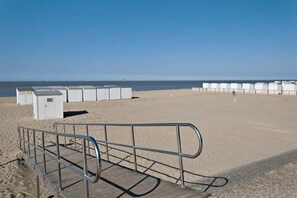 A beach with a wooden walkway and metal railing, leading to white beach huts. - Cozy apartment w/ balcony & parking (Knokke-Heist)