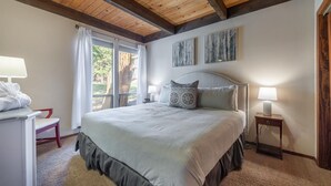 A bedroom with a large bed, two bedside tables with lamps, a window with curtains, and a view of trees outside. - Kingswood Village 57 3 Bedroom Condo (Kings Beach)