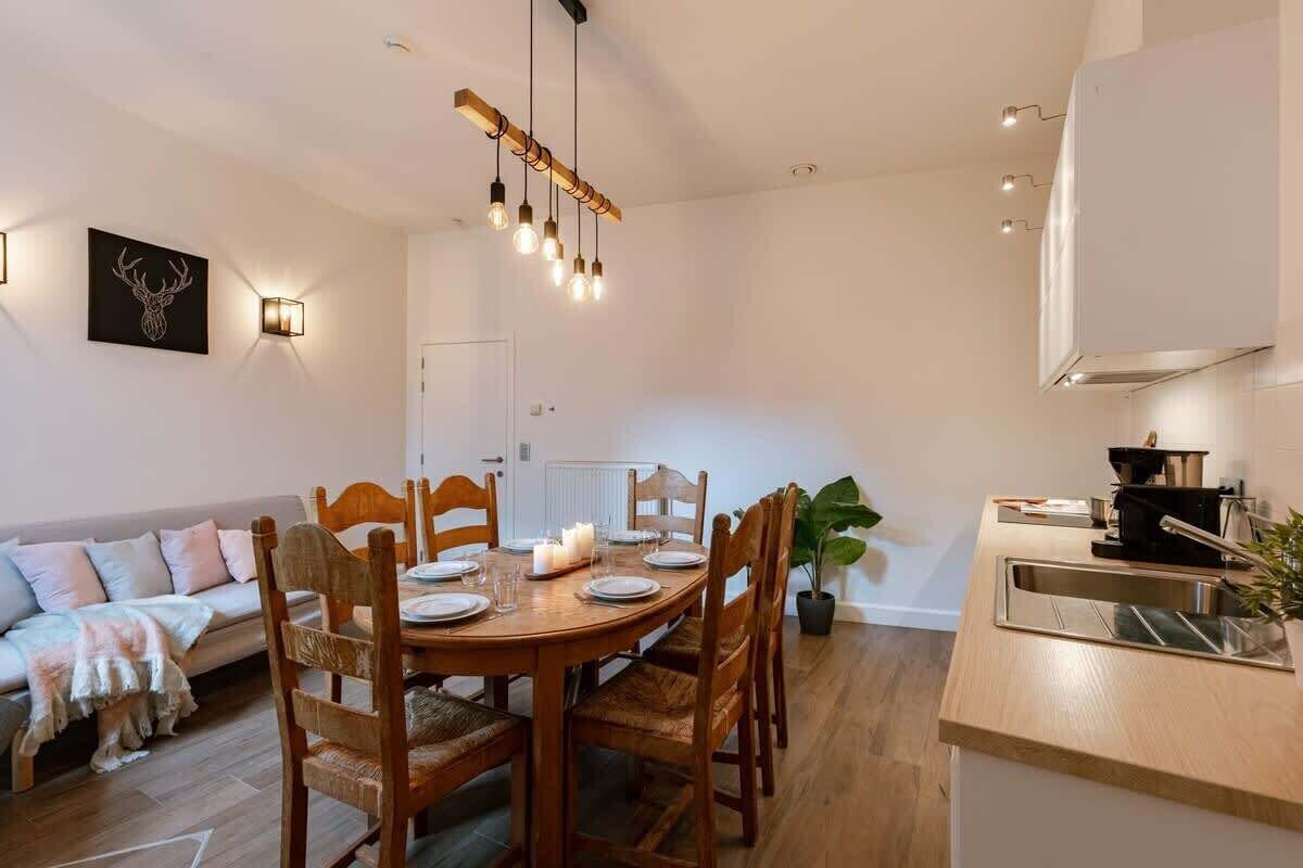 Een eetruimte met een houten tafel voor vier personen, omringd door houten stoelen. Aan de rechterkant is een keuken met een spoelbak en fornuis.