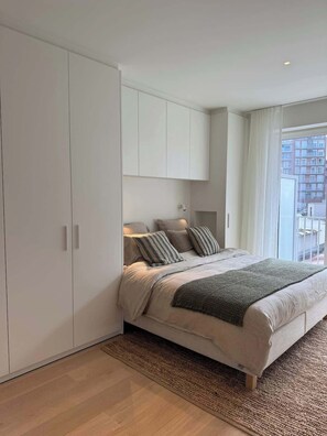 Un dormitorio con cama, armario, ventana con cortinas y vista a edificios.