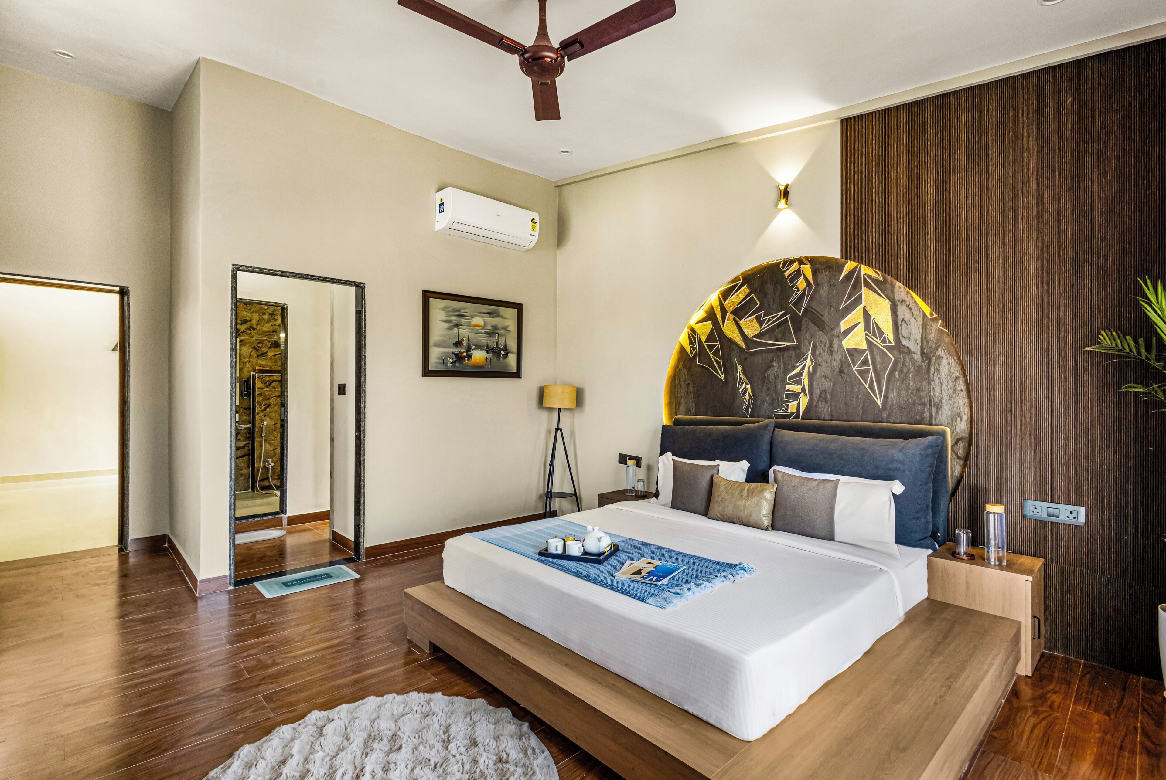 Una habitación de hotel moderna con una cama grande, un ventilador de techo, suelo de madera, una mesita de noche y un aire acondicionado instalado en la pared.