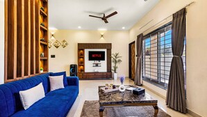 A living room with a blue sofa, a coffee table, a TV, and a large window with curtains. - ELIVAAS Casa llios Stuning Getaway Villa (Udaipur)