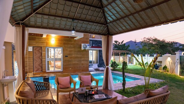 A covered outdoor seating area with wicker furniture, a pool, and a view of the surrounding landscape. - ELIVAAS Casa llios Stuning Getaway Villa (Udaipur)