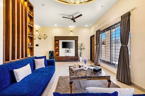 A modern living room with a blue sofa, a coffee table, a TV, and a ceiling fan. - ELIVAAS Casa llios Stuning Getaway Villa (Udaipur)