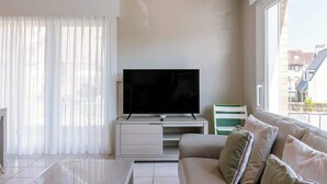 Uma sala de estar moderna com uma televisão de tela plana em uma unidade de entretenimento branca, um sofá com almofadas e uma grande janela com persianas.
