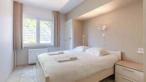 Um quarto com cama, mesas de cabeceira, radiador e janela com cortinas.