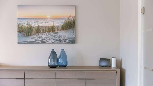 Um quadro pendurado na parede retratando uma cena de praia, um armário de madeira com dois vasos e um pequeno alto-falante.