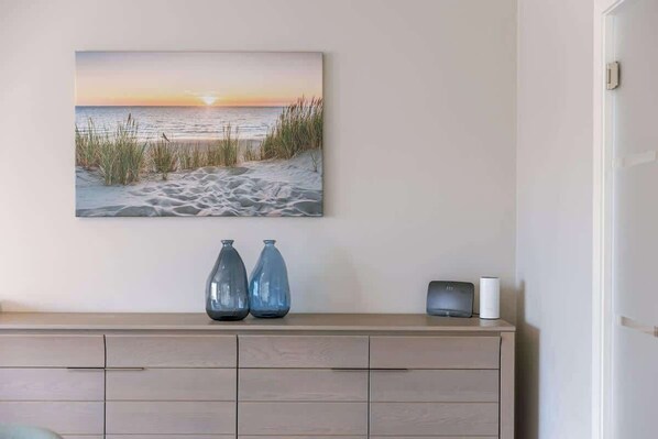 Un tableau représentant une scène de plage, accroché au mur, un meuble en bois avec deux vases et une petite enceinte.
