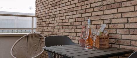 A balcony with a brick wall, a wooden table, two chairs, and a wicker basket with a bottle and glasses.