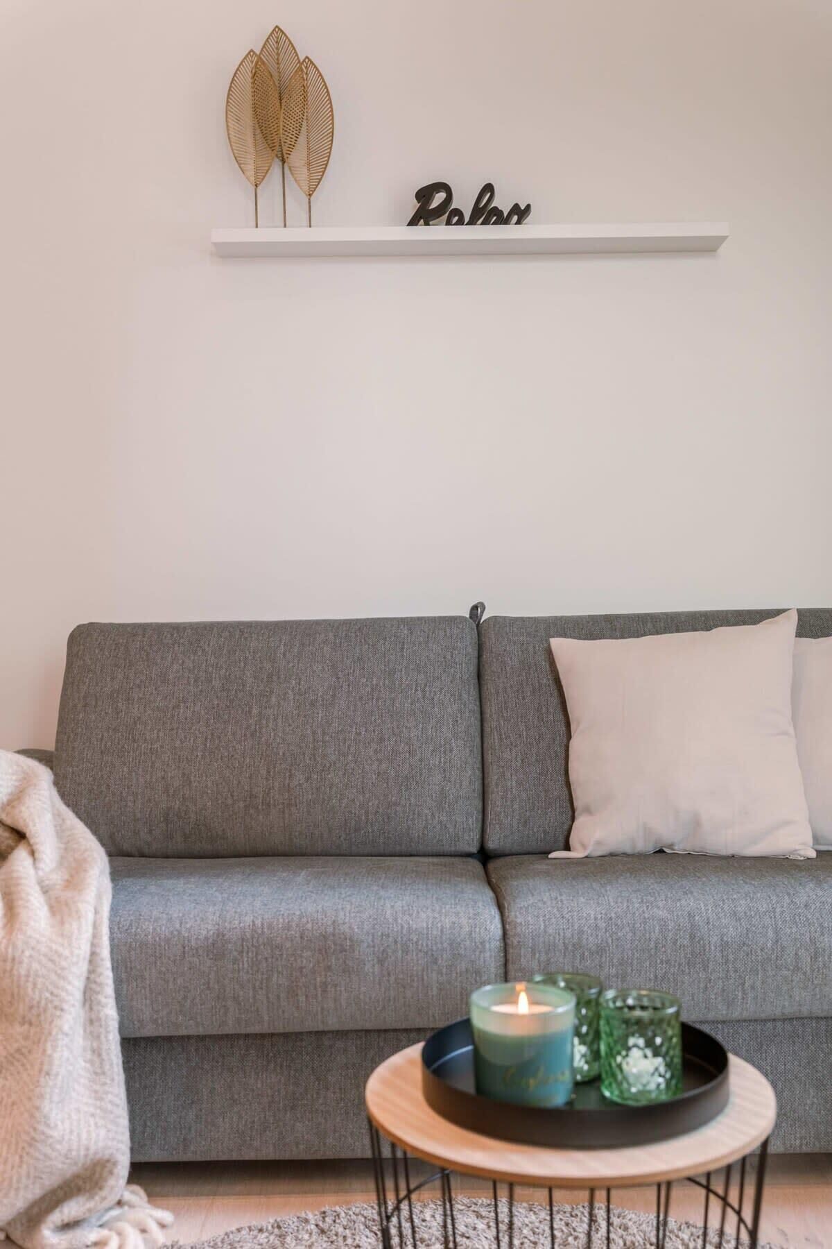 Un sofá gris con un cojín blanco, una estantería con hojas decorativas y la palabra 'Relax', una pequeña mesa redonda con una vela y recipientes de vidrio verde, y una manta gris en el suelo.
