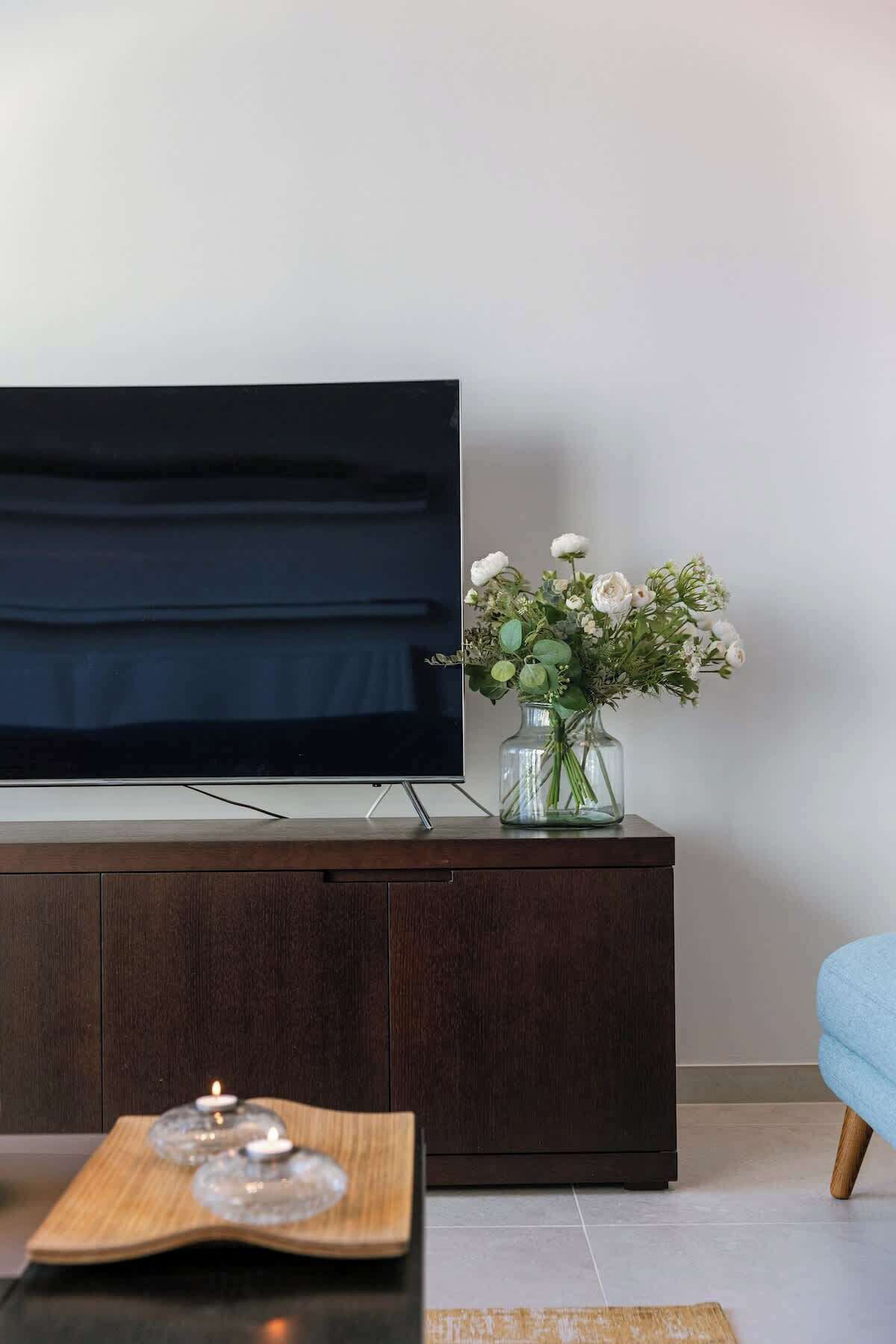 Uma televisão de tela plana sobre um móvel de madeira, um vaso com flores e um castiçal sobre uma mesa.