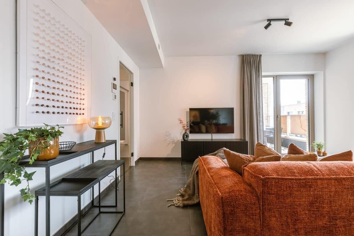 A modern living room with a sofa, a TV, a wall-mounted light fixture, and a large window.