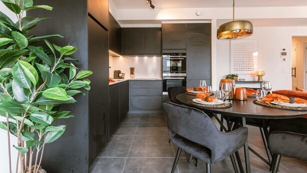 Una cocina moderna con muebles oscuros, un comedor con una mesa redonda y sillas, y una planta grande en maceta.