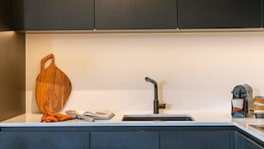 Una cocina moderna con armarios oscuros, una encimera blanca, una tabla de cortar de madera, un lavamanos con grifo y una cafetera.