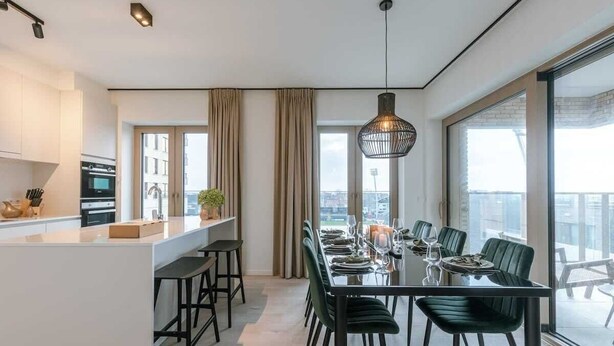 Una cocina moderna con una isla central, un comedor con mesa y sillas de vidrio, y grandes ventanas que ofrecen una vista.