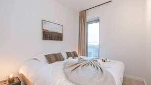Una habitación de hotel moderna con una cama grande, una mesita de noche, una lámpara de noche, una ventana con cortinas y una obra de arte colgada en la pared.