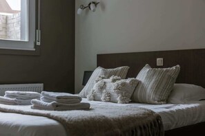 Een opgemaakt bed met witte lakens en zachte kussens, een raam met uitzicht op een gebouw en een wandlamp.