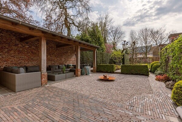 A covered outdoor seating area with brick walls, a fire pit, and neatly trimmed hedges.