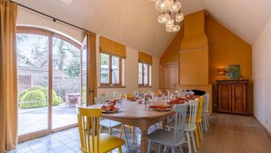 Ein Essbereich mit einem großen Tisch, gedeckt für sechs Personen, umgeben von Stühlen, ein Küchenbereich mit Holzschränken und Blick durch eine Glastür auf eine Terrasse.