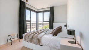 A modern bedroom with a large window, a bed with a white headboard, a bedside table with a lamp, and a bench by the window. - Bright 2BR apartment w/ balcony & private entrance (Gent)