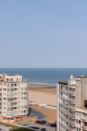A coastal view with high-rise buildings, a beach, and a road with parked cars. - 1BR unit w/ parking & elevator (Oostende)