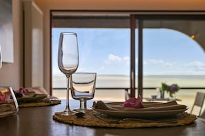 Une table à manger offrant une vue dégagée sur la mer grâce à une grande fenêtre, un verre d’eau et une assiette ornée d’une fleur.
