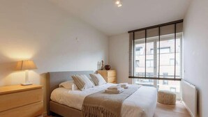 A bedroom with a bed, bedside table, lamp, window, and radiator. - Beachfront Gem w/ Balcony & Family Friendly in (Knokke-Heist)