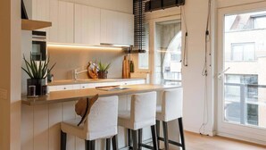A modern kitchen with white cabinets, a central island with bar stools, a window with blinds, and a visible outdoor view. - Beachfront Gem w/ Balcony & Family Friendly in (Knokke-Heist)