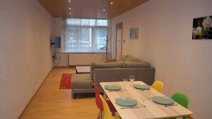 A dining area with a table set for four, a grey sofa, and a window with a view of parked cars. - Beachfront Apartment w/ Patio, Pet-Friendly, in (Oostende)