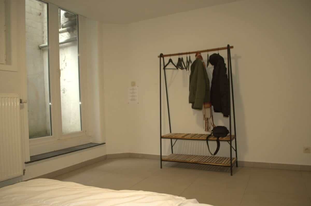 Une pièce avec un lit, une fenêtre, un radiateur et un portant à vêtements avec des vestes.