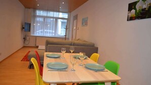 A dining area with a table set for four, a sofa in the background, and a window with blinds. - Beachfront Apartment w/ Patio, Pet-Friendly, in (Oostende)