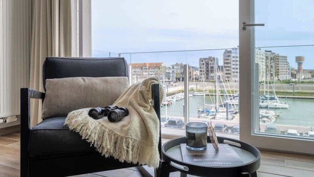 A modern room with a black leather chair, a blanket, and a camera on the armrest. A small table with a candle and a glass is in front of a large window overlooking a marina with boats and buildings. - Waterfront studio w/ private entrance (Blankenberge)