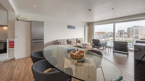 A modern living space with a glass dining table, a sofa set, and a view of the city through large windows. - Waterfront studio w/ private entrance (Blankenberge)