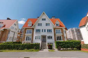 Ein mehrgeschossiges Wohngebäude mit einer symmetrischen Fassade, einem zentralen Eingang mit Treppe und einem roten Ziegeldach.