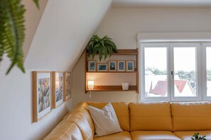 Ein Wohnzimmer mit einem gelben Sofa, einem Holzbodenregal mit Bildern und einer Pflanze sowie einem Fenster mit Blick auf Gebäude.