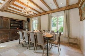 A dining area with a wooden table and chairs, a chandelier, and a view of greenery outside.