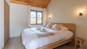 A bedroom with a wooden ceiling, a bed with white bedding, a window with curtains, and a small stool.