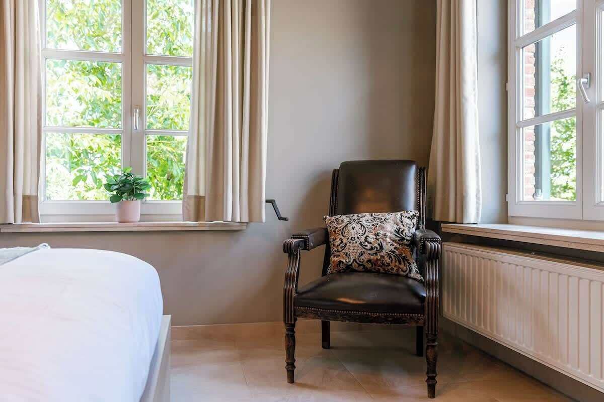 Una habitación con una cama, una ventana con cortinas, un sillón con un cojín estampado y una planta en maceta en el alféizar.