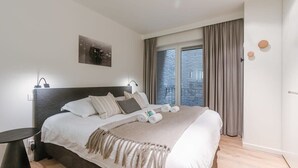 A neatly made bed with pillows and a blanket, a bedside table, a wall-mounted light, a window with curtains, and a small wall decoration. - Casiola coastal stay - parking & terrace (Knokke-Heist)